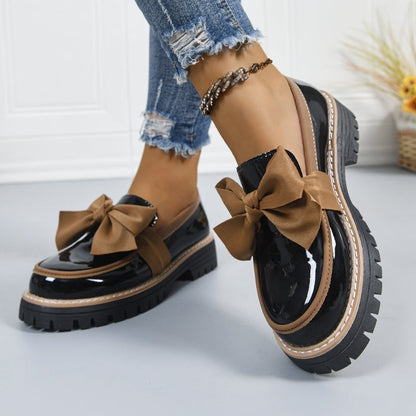 Mocasines con lazo marrón sobre cuero negro, suela gruesa y borde claro.