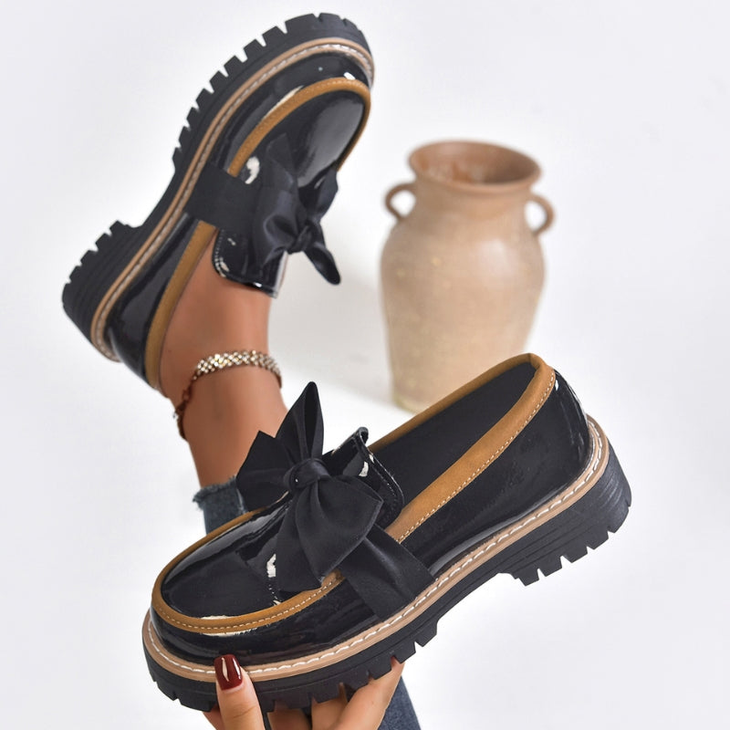 Mocasines con lazo negro, empeine brillante y suela gruesa con ribete claro.
