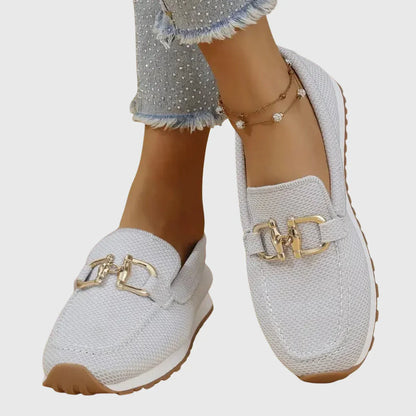 Zapatillas tipo mocasín Chiara blancas con adorno metálico dorado y costuras visibles.